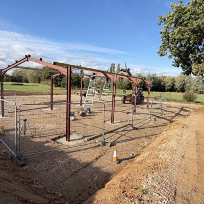 The red steel frame of the new agricultural barn going up on a stone yard at Bullbanks Farm, with fields and trees behind