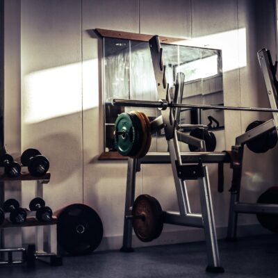 An empty gym in soft light with a loaded barbell on a squat rack and a row of dumbbells beside it
