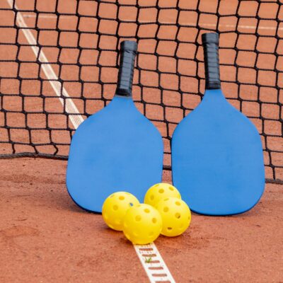 Two blue pickleball paddles leaning against a net with four yellow pickleballs on the court in front of them