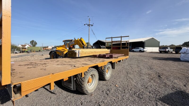 A farm trailer loaded with timber and red steel sections for the new agricultural barn, with a telehandler and farm buildings behind