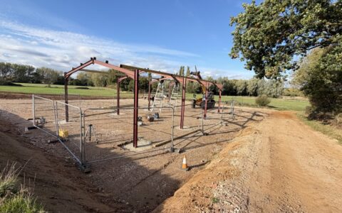 The red steel frame of the new agricultural barn going up on a stone yard at Bullbanks Farm, with fields and trees behind