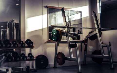 An empty gym in soft light with a loaded barbell on a squat rack and a row of dumbbells beside it