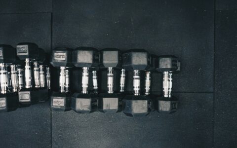 A row of black hex dumbbells with silver handles lined up in size order on a dark rubber gym floor